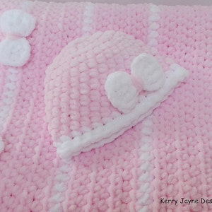 K&ouml;nnte beinhalten: Eine rosa und wei&szlig;e geh&auml;kelte Babydecke mit einer passenden geh&auml;kelten M&uuml;tze. Die Decke hat wei&szlig;e Streifen und wei&szlig;e Bommeln. Die M&uuml;tze hat eine wei&szlig;e Schleife.
