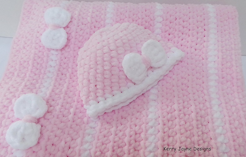 K&ouml;nnte beinhalten: Eine rosa und wei&szlig;e geh&auml;kelte Babydecke mit einer passenden geh&auml;kelten M&uuml;tze. Die Decke hat wei&szlig;e Streifen und wei&szlig;e Bommeln. Die M&uuml;tze hat eine wei&szlig;e Schleife.