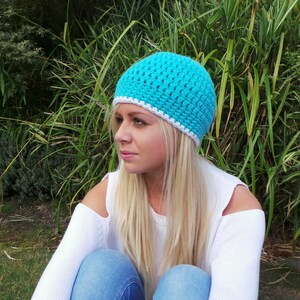 May include: A turquoise crocheted beanie with a white trim. The beanie is worn by a person with long blonde hair. The person is sitting in a grassy area.