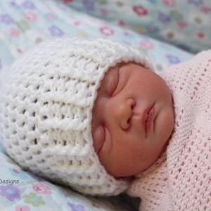 May include: A white crocheted baby hat with a textured pattern. The hat is on a baby sleeping on a floral patterned blanket.