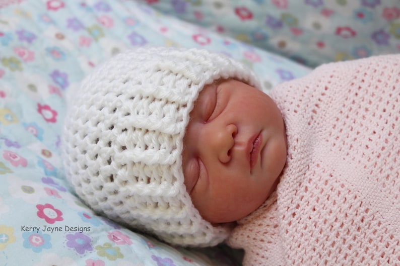 May include: A white crocheted baby hat with a textured pattern. The hat is on a baby sleeping on a floral patterned blanket.