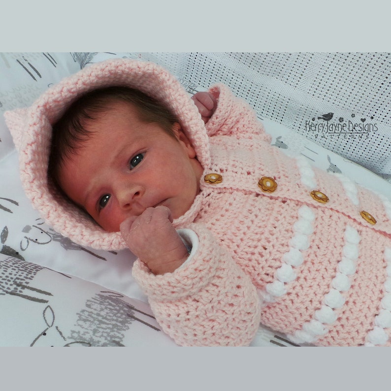 May include: A pink crocheted baby cardigan with a hood and white accents. The cardigan has wooden buttons and is worn by a baby.