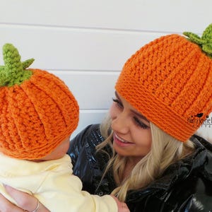 May include: A woman and a baby wearing matching orange and green crocheted pumpkin hats. The hats have a textured stitch pattern and a green stem on top.