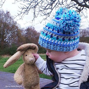 May include: A child wearing a blue, white, and gray striped crocheted hat with a large blue pom-pom on top. The child is holding a brown plush bunny rabbit.