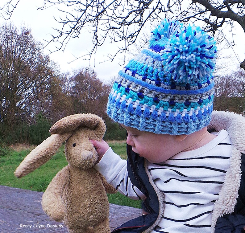 May include: A child wearing a blue, white, and gray striped crocheted hat with a large blue pom-pom on top. The child is holding a brown plush bunny rabbit.