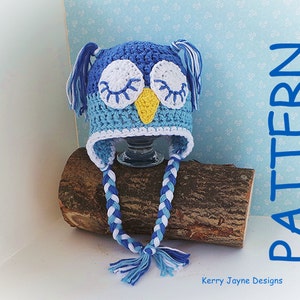 May include: Blue and white crocheted owl hat with long braided ties. The hat has a yellow beak and white eyes. The hat is sitting on a piece of wood. "PATTERN" is written vertically in blue on the right side of the image. "Kerry Jayne Designs" is written in black at the bottom of the image.