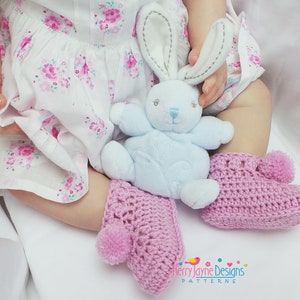 Puede incluir: Un par de botitas de beb&eacute; de crochet rosa con un pomp&oacute;n en cada dedo del pie. Las botitas las lleva un beb&eacute; sentado en una superficie blanca, sosteniendo un conejito de peluche azul.