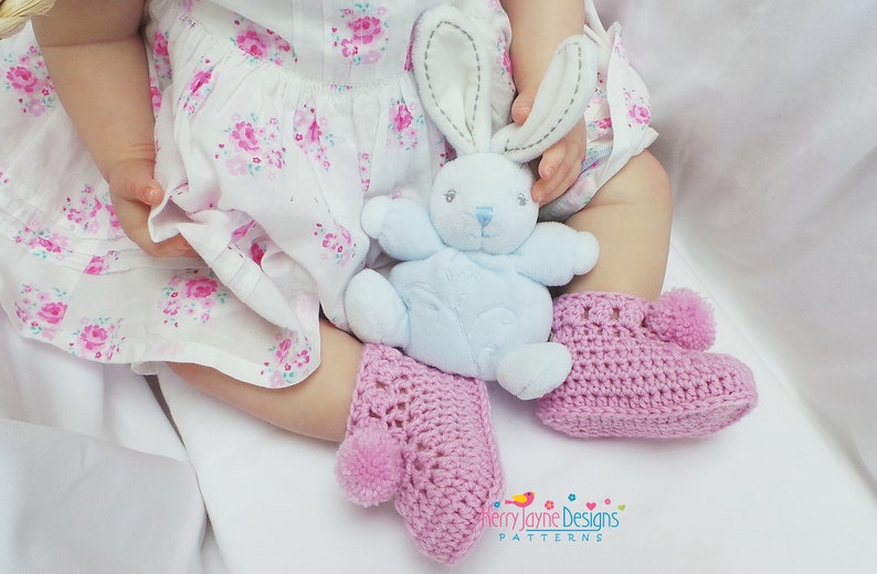 Puede incluir: Un par de botitas de beb&eacute; de crochet rosa con un pomp&oacute;n en cada dedo del pie. Las botitas las lleva un beb&eacute; sentado en una superficie blanca, sosteniendo un conejito de peluche azul.