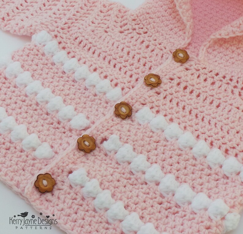 May include: A pink crocheted baby cardigan with white bobbles and wooden flower buttons.