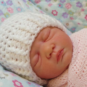 May include: A white crocheted baby hat with a textured pattern. The hat is on a baby sleeping on a floral patterned blanket.