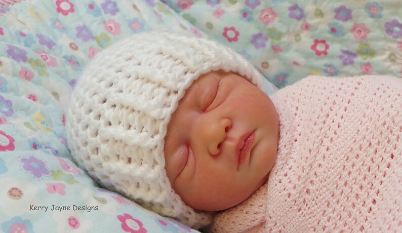 May include: A white crocheted baby hat with a textured pattern. The hat is on a baby sleeping on a floral patterned blanket.