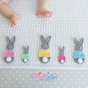 May include: A white crocheted baby blanket with a row of grey bunny appliques. The bunnies are wearing different colored crocheted outfits, including pink, yellow, green, and blue. The blanket has a white pom-pom on one corner. "Kerry Jayne Designs Patterns" is written on the blanket.