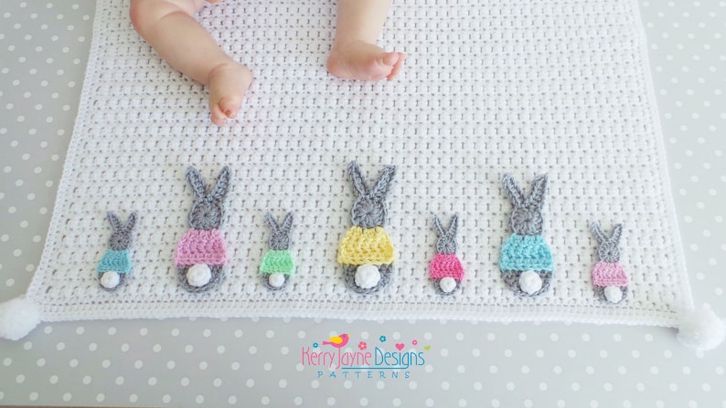 May include: A white crocheted baby blanket with a row of grey bunny appliques. The bunnies are wearing different colored crocheted outfits, including pink, yellow, green, and blue. The blanket has a white pom-pom on one corner. "Kerry Jayne Designs Patterns" is written on the blanket.