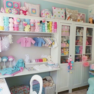 May include: A white shelf unit filled with colorful yarn, crocheted items, and stuffed animals. The shelf unit is in a room with a light blue wall and a white desk with a white chair. The desk has a sign that says "Homemade Designs Patterns".
