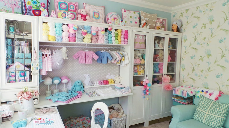 May include: A white shelf unit filled with colorful yarn, crocheted items, and stuffed animals. The shelf unit is in a room with a light blue wall and a white desk with a white chair. The desk has a sign that says "Homemade Designs Patterns".
