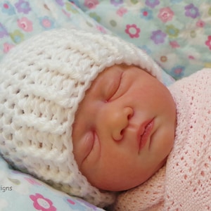 May include: A white crocheted baby hat with a textured pattern. The hat is on a baby sleeping on a floral patterned blanket.