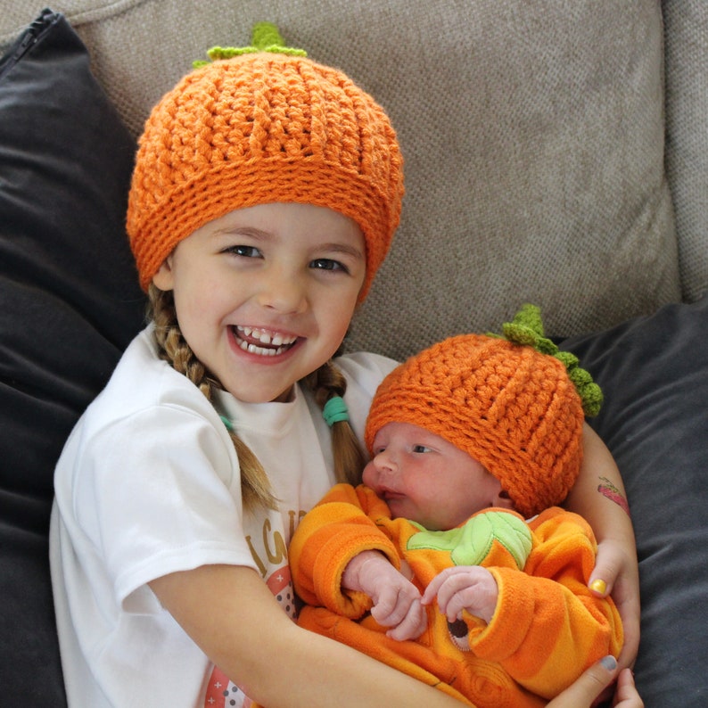 May include: A young girl wearing an orange crocheted pumpkin hat holds a baby wearing an orange crocheted pumpkin hat and an orange pumpkin-shaped outfit.