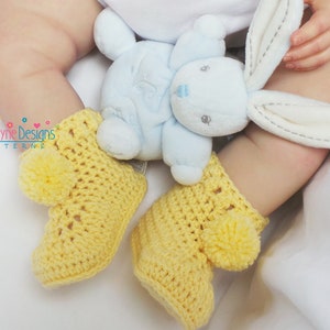 Puede incluir: Un par de botitas de beb&eacute; de crochet amarillo con pompones en la parte superior. Las botitas las lleva un beb&eacute; que sostiene un conejito de peluche azul.