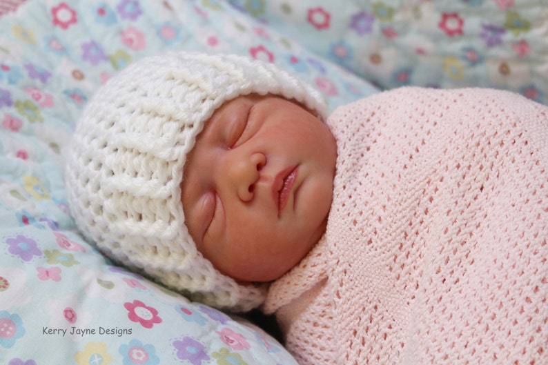 May include: A newborn baby wearing a white crocheted hat and a pink crocheted blanket. The baby is sleeping peacefully on a floral patterned fabric.