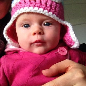 May include: A baby wearing a pink and white crocheted owl hat with a yellow beak and blue accents. The baby is wearing a pink quilted jacket with a white button.