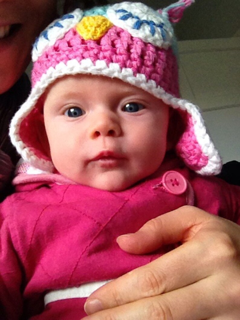May include: A baby wearing a pink and white crocheted owl hat with a yellow beak and blue accents. The baby is wearing a pink quilted jacket with a white button.