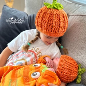 May include: Two children wearing crocheted pumpkin hats. The older child is wearing a white shirt with the word "Sister" on it. The younger child is wearing an orange pumpkin costume.
