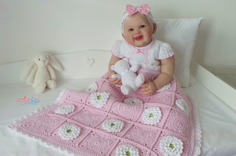 May include: A pink crocheted baby blanket with white flowers. The blanket has a white crocheted border. A baby doll is sitting on the blanket holding a white teddy bear.