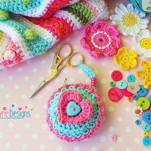 May include: A crocheted heart-shaped measuring tape with a blue and pink flower design. The tape is surrounded by colorful buttons, yarn, and crocheted flowers. The text "Kerry Jayne Designs" is visible in the lower left corner of the image.