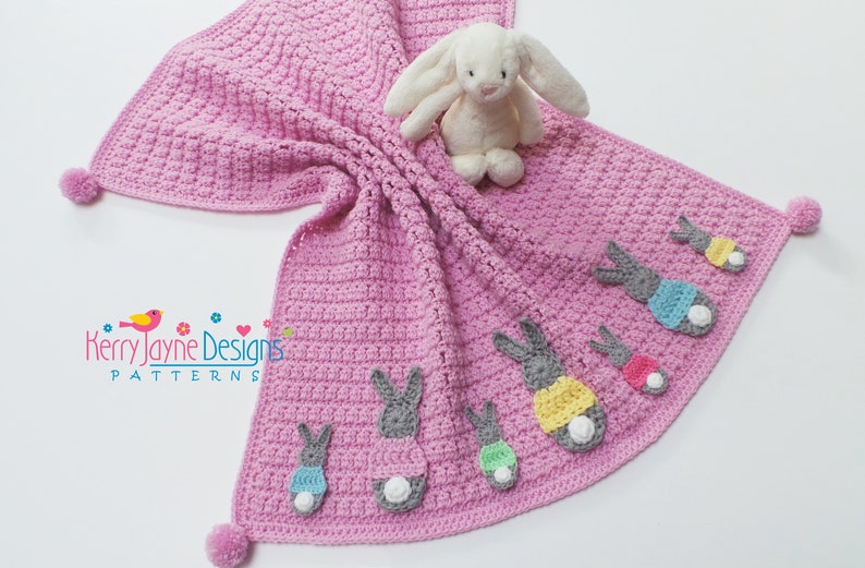 May include: A pink crocheted baby blanket with a white bunny and several colorful bunny appliques. The blanket has a pom-pom on each corner.