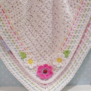 Peut inclure: Une couverture pour b&eacute;b&eacute; en crochet blanc avec une bordure color&eacute;e et des appliques de fleurs. La couverture a un fond bleu clair avec des pois blancs.