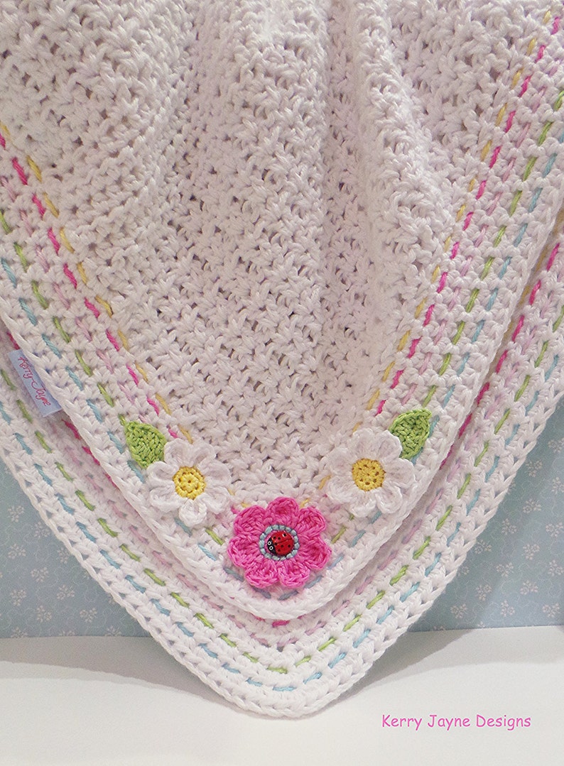 Peut inclure: Une couverture pour b&eacute;b&eacute; en crochet blanc avec une bordure color&eacute;e et des appliques de fleurs. La couverture a un fond bleu clair avec des pois blancs.