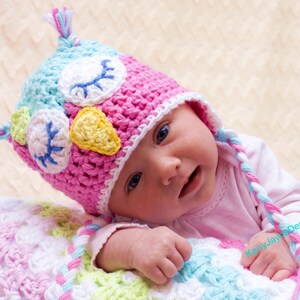 May include: A baby wearing a pink and blue crocheted owl hat with a white and yellow beak. The hat has a long, braided strap that is tied under the chin. The baby is lying on a white and pink crocheted blanket.