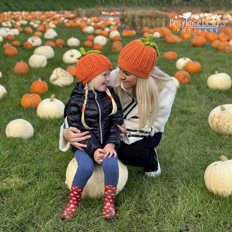 May include: A mother and daughter wearing orange pumpkin-shaped crocheted hats in a field of pumpkins. The girl is sitting on a large white pumpkin and the woman is standing behind her. The text "Kerry Jayne Designs PATTERNS" is visible in the top right corner of the image.