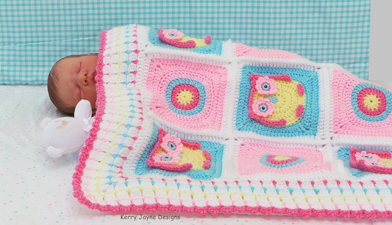 May include: A pink, blue, and white crocheted baby blanket with a border of pink and white. The blanket features a repeating pattern of owls and circles. The blanket is folded over a baby doll.