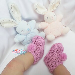 Puede incluir: Un par de botitas de beb&eacute; de crochet rosa con pompones en los dedos. Las botitas est&aacute;n en los pies de un beb&eacute; que lleva ropa blanca. Dos juguetes de peluche de conejo est&aacute;n en el fondo.