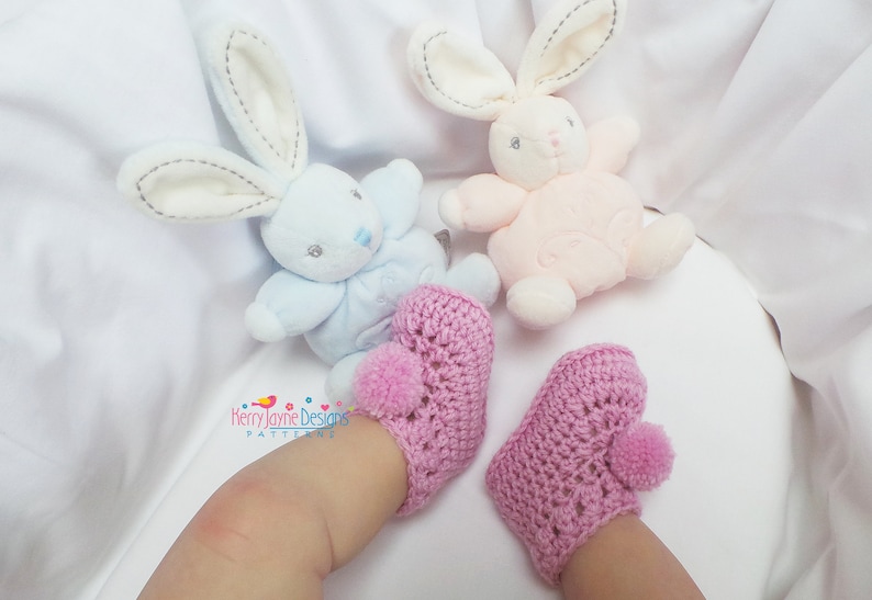 Puede incluir: Un par de botitas de beb&eacute; de crochet rosa con pompones en los dedos. Las botitas est&aacute;n en los pies de un beb&eacute; que lleva ropa blanca. Dos juguetes de peluche de conejo est&aacute;n en el fondo.