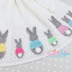 May include: A white crocheted baby blanket featuring several gray bunny figures with colorful accents. The bunnies have pink, yellow, and blue clothing and white fluffy tails. The blanket has a pom-pom trim and a pom-pom at the corner.