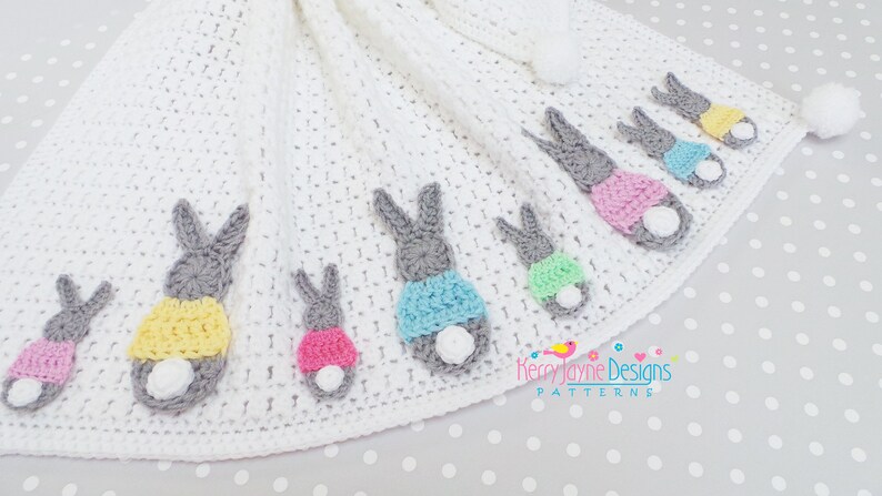 May include: A white crocheted baby blanket featuring several gray bunny figures with colorful accents. The bunnies have pink, yellow, and blue clothing and white fluffy tails. The blanket has a pom-pom trim and a pom-pom at the corner.