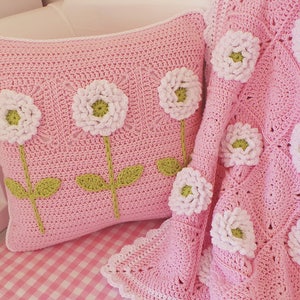 May include: A pink crocheted pillow and blanket with white flowers and green stems. The pillow has four flowers and the blanket has several flowers.