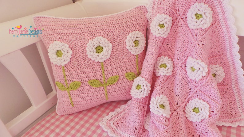 May include: A pink crocheted pillow and blanket with white flowers and green stems. The pillow has four flowers and the blanket has several flowers.