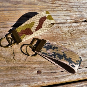 Pode incluir: Dois chaveiros com tecido de camuflagem e argolas de chave de latão. Um chaveiro tem um padrão de camuflagem marrom, verde e bege. O outro chaveiro tem um padrão de camuflagem rosa, marrom e preto.