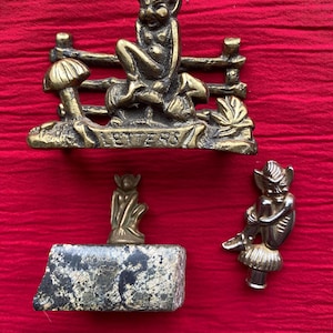 Könnte beinhalten: Drei Messing-Feenfiguren auf rotem Hintergrund. Eine Fee sitzt auf einem Pilz und eine andere auf einer Bank mit dem Wort "LETTERS". Die dritte Fee sitzt auf einem Stein.