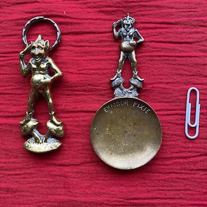 May include: Two brass Exmoor Pixie figurines. One is a keychain, the other is a spoon. Both feature a standing pixie figure with mushroom-shaped feet. The spoon has the words "Exmoor Pixie" engraved on it. A white paperclip is next to the figurines.