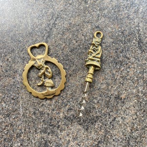 May include: A set of two brass bottle openers featuring a whimsical design of a fairy sitting on a mushroom. The openers are both gold in color and have a detailed design.