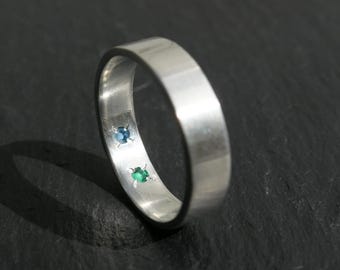 May include: A silver ring with a flat band design. The ring features two small gemstones, one blue and one green, set into the band. The ring is displayed on a dark, textured surface, highlighting its polished metal finish.