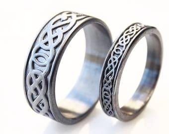 May include: Two silver wedding bands with a Celtic knot design. The bands are both wide and have a smooth finish.