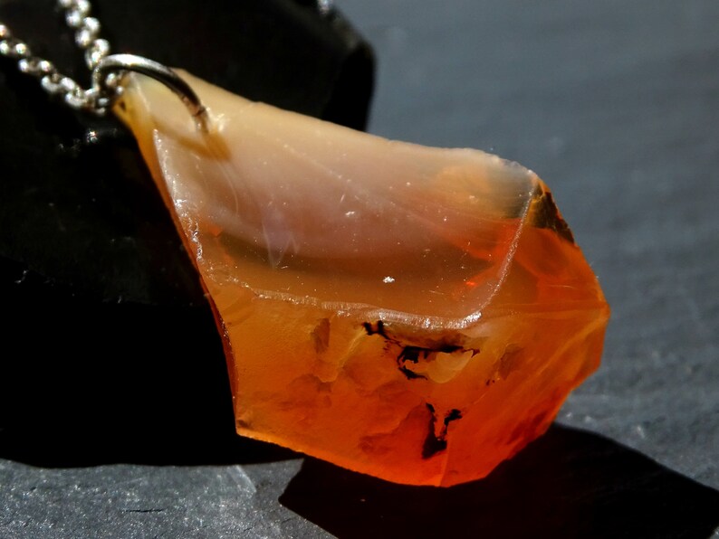 May include: A pendant necklace featuring a large, irregularly shaped orange and clear gemstone. The gemstone has a rough, natural texture and is attached to a silver chain.
