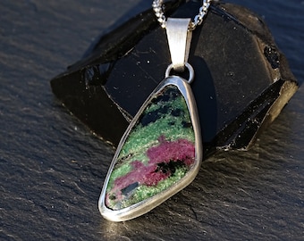 May include: A silver pendant with a green and pink gemstone set in a silver bezel. The pendant is on a black background.