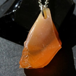 May include: A pendant necklace featuring a raw, orange-colored opal stone. The stone is irregularly shaped and has a translucent quality. The pendant is attached to a silver chain.