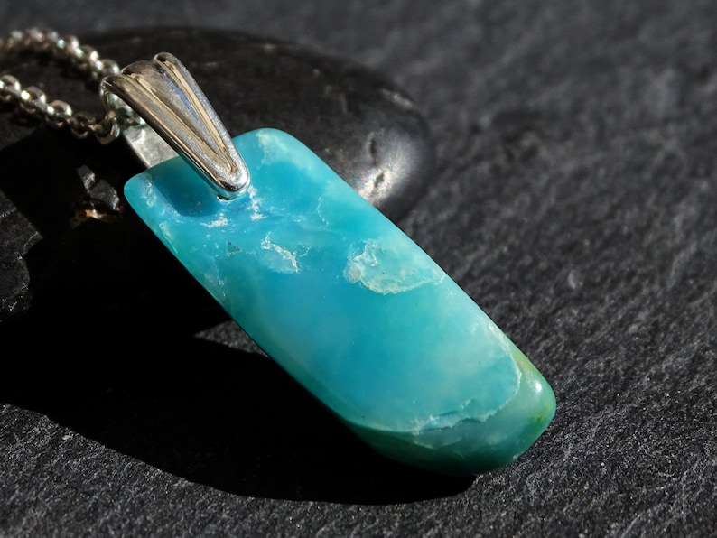 May include: A turquoise blue gemstone pendant with a silver bail. The pendant is on a black surface.